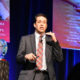 foster parent: Man on stage in front of 2 large banners, wearing suit and tie, gestures.