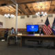 Syracuse: 2 men in suits sit at table, one woman stands near 2 flags