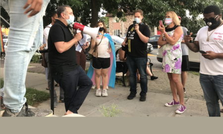 Schenectady: Man in black with megaphone wearing mask talks to small group wearing masks, holding up phones. One woman is wearing superhero-style cape.