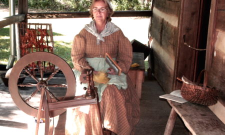 Woman in long dress, kerchief sits before spinning wheel on wooden porch
