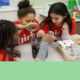 Nonprofit COVID Relief: Young girls in red t-shirts crowd aroun table looking at items through a magnifying glass