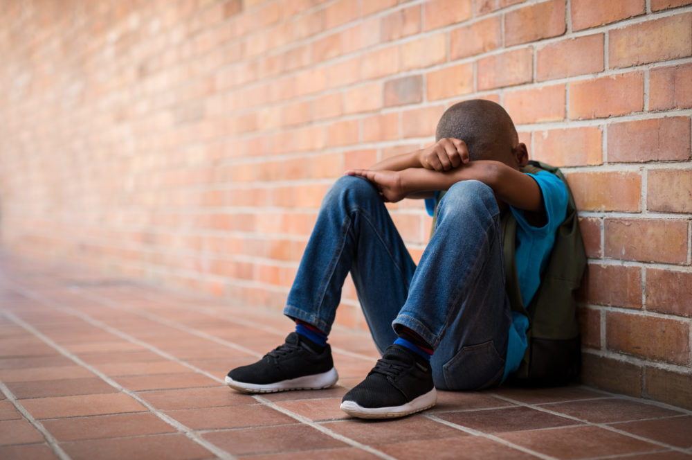 Black children investigated by CPS: sad black child sitting against brick wall with head in arms