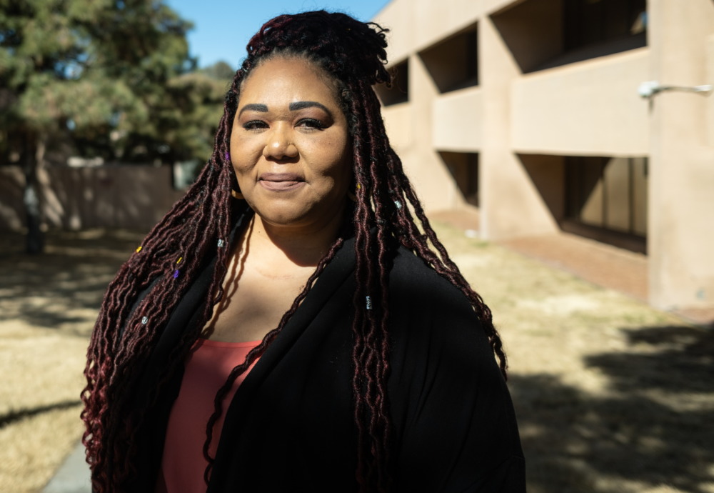 New Mexico Sex Trafficking: Middle-aged woman with long dark hair wearing black jacket with red blouse stands outside in frone of a beige stucco building and trees.