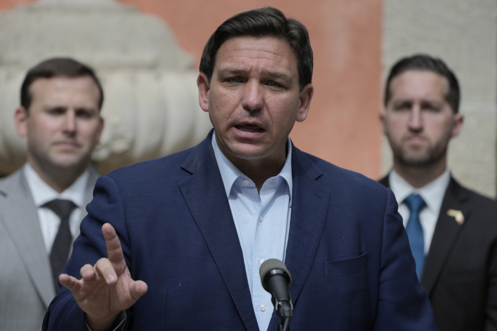 Don't Say Gay Florida: closeup of a white man in a blue sut gestering and speaking into a microphone flanked by two white men in suits