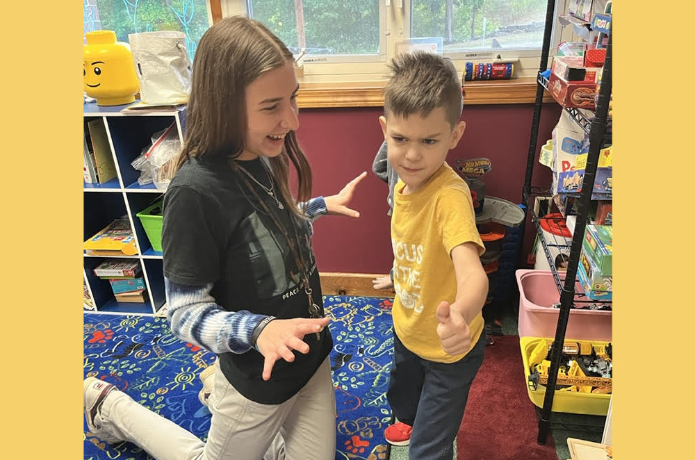 Microschool: Young woman with long, dark hair in nave top and white pants kneels on floor with young boy in yellow shirt and jeans standing next to her.