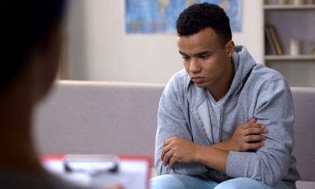 at-risk youth, Community-youth-violence-prevention: Black teen sullenly sits on couch across from counselor