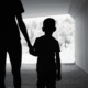 foster care: Black and white photo of silhouette back of adult holding hand of young child, walking away from camera in dark tunnel toward light outdoor area