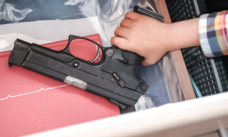 Yout gun deaths: Very young child's hand and arm in red plaid shirt reaches into drawer grabbing a large black gun