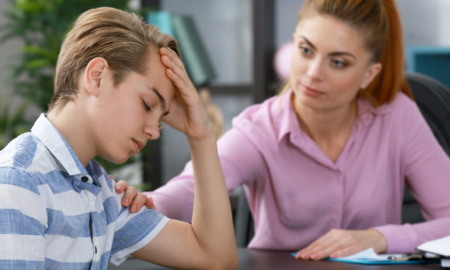Research boys expressing feelings: Young teen boy in blue and white striped shirt sits at desk looking down with head in hand while sitting across from to an adult female in pink shirt speaking to him